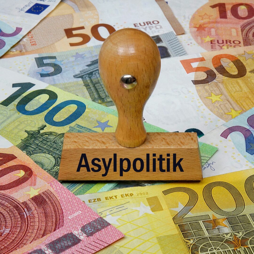 Auf dem Bild ist ein Stempel mit der Aufschrift "Asylpolitik" zu sehen, darunter liegen mehrere Geldscheine.