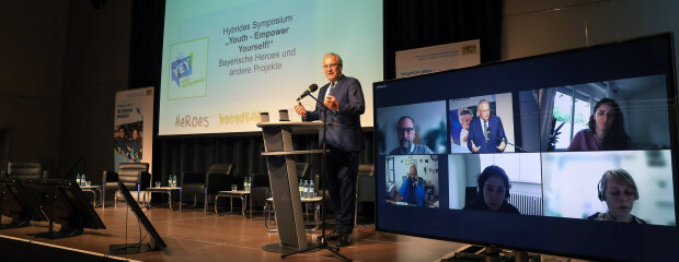 Innen- und Integrationsminister Joachim Herrmann hinter Rednerpult neben Bildschirm mit Videokonferenz